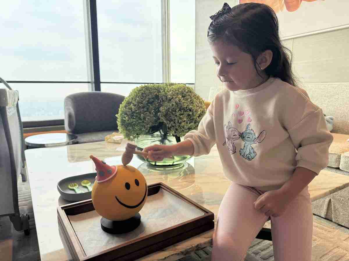 A young girl taps a smiling yellow ball with a small mallet while sitting at a table in a modern living room.