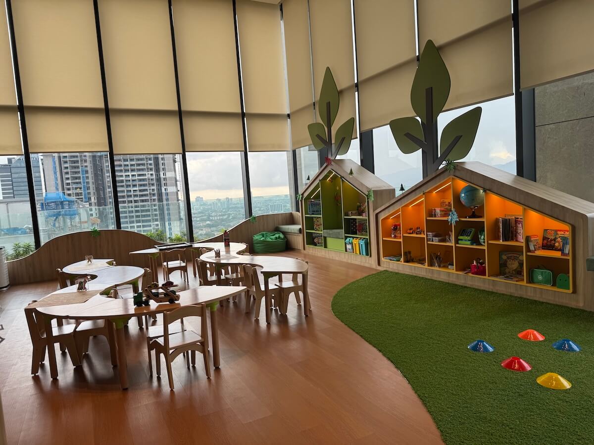 A children's playroom with wooden tables and chairs, playhouses with shelves of toys and books, artificial grass, and city views from large windows.