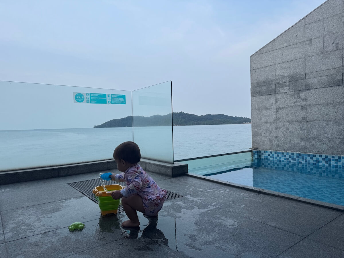 A toddler in patterned clothing plays with toys near an outdoor pool, beside a glass barrier overlooking the ocean and a distant hilly shoreline.