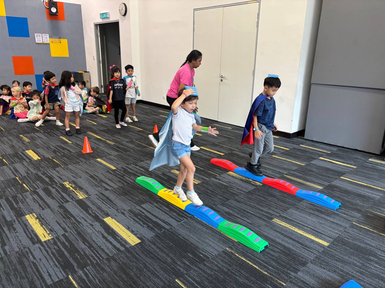 Children participate in a balancing activity indoors, stepping on colorful beams, while others wait in line. An adult supervises the activity.