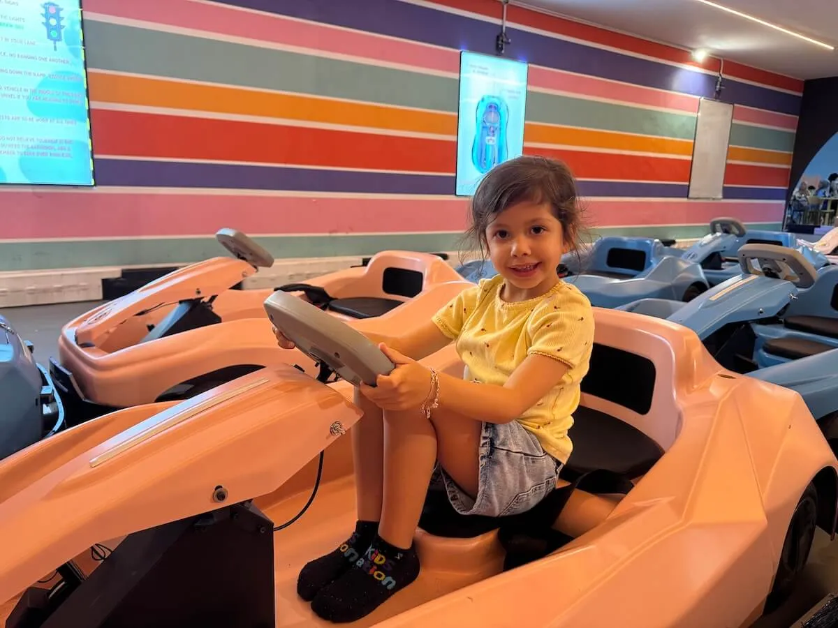 A young girl sits in a peach-colored toy car at an indoor play area with colorful striped walls in the background.