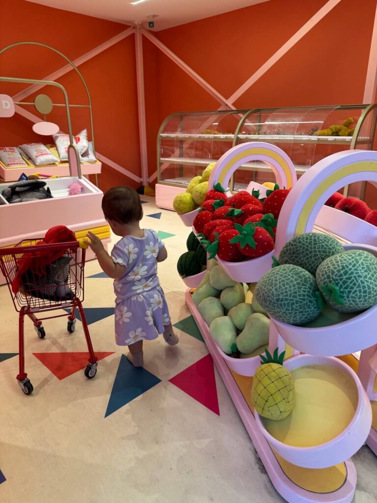 A small child in a purple outfit pushes a toy shopping cart near shelves of large plush fruit in a brightly colored play grocery store.
