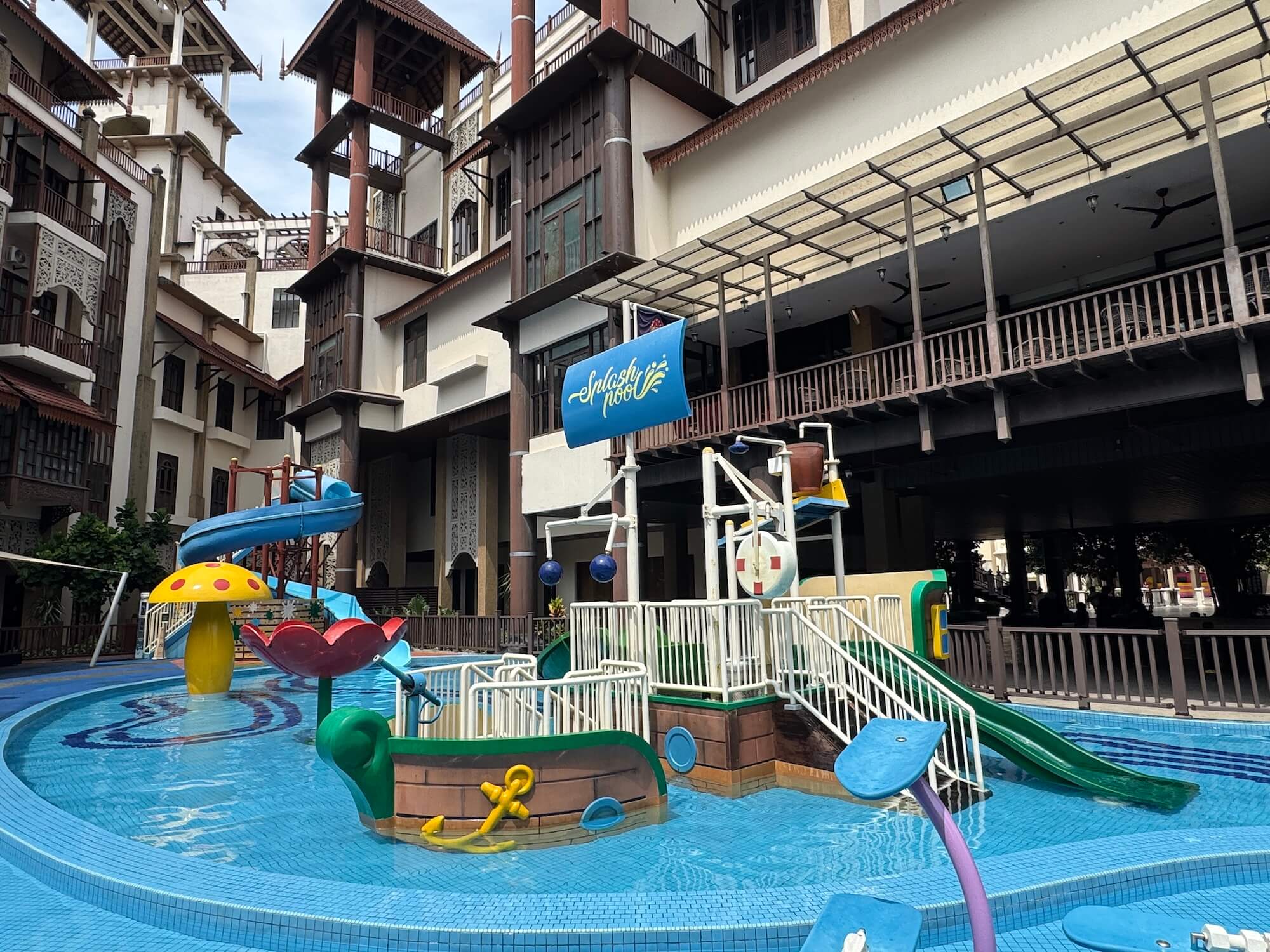 Outdoor children's water play area with slides, climbing structures, and splash features, surrounded by multi-story buildings with balconies and shaded walkways.